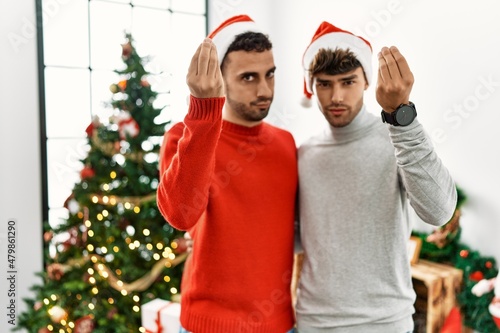 Young gay couple standing by christmas tree wearing hat doing italian gesture with hand and fingers confident expression