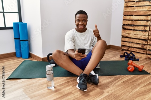 Young african man sitting on training mat at the gym using smartphone doing happy thumbs up gesture with hand. approving expression looking at the camera showing success.
