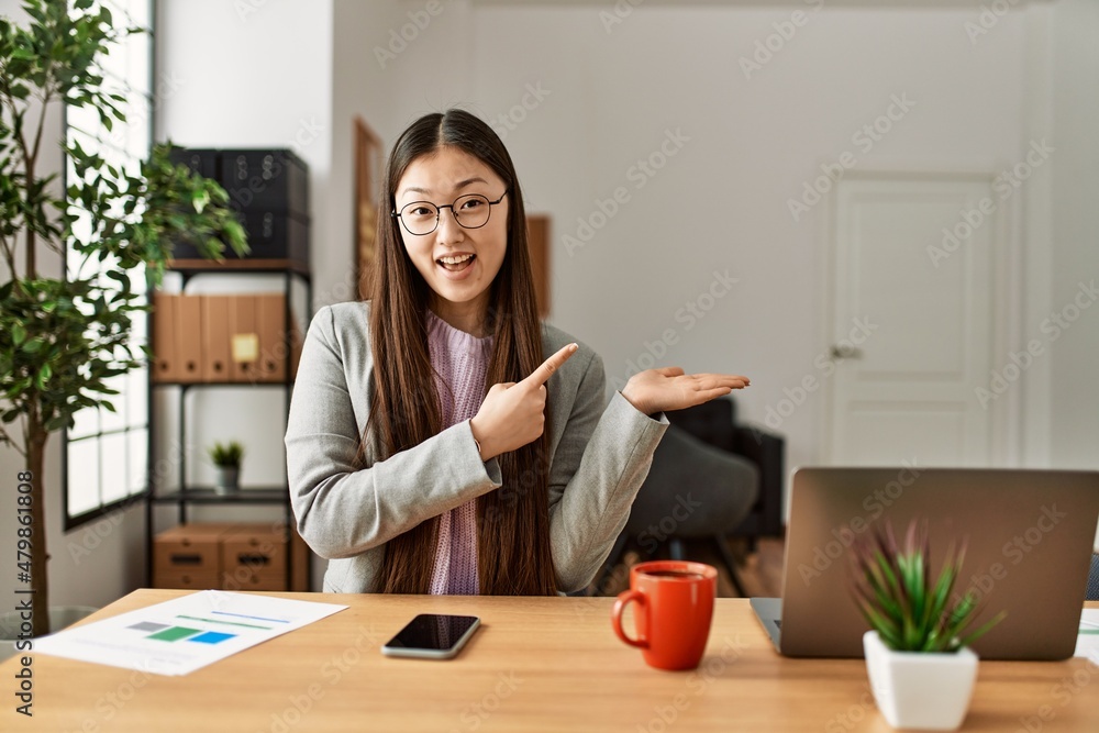 Young chinese business worker wearing business style sitting on desk at office amazed and smiling to the camera while presenting with hand and pointing with finger. Young chinese business worker wearing business style sitting on desk at office amazed and smiling to the camera while presenting with hand and pointing with finger.
