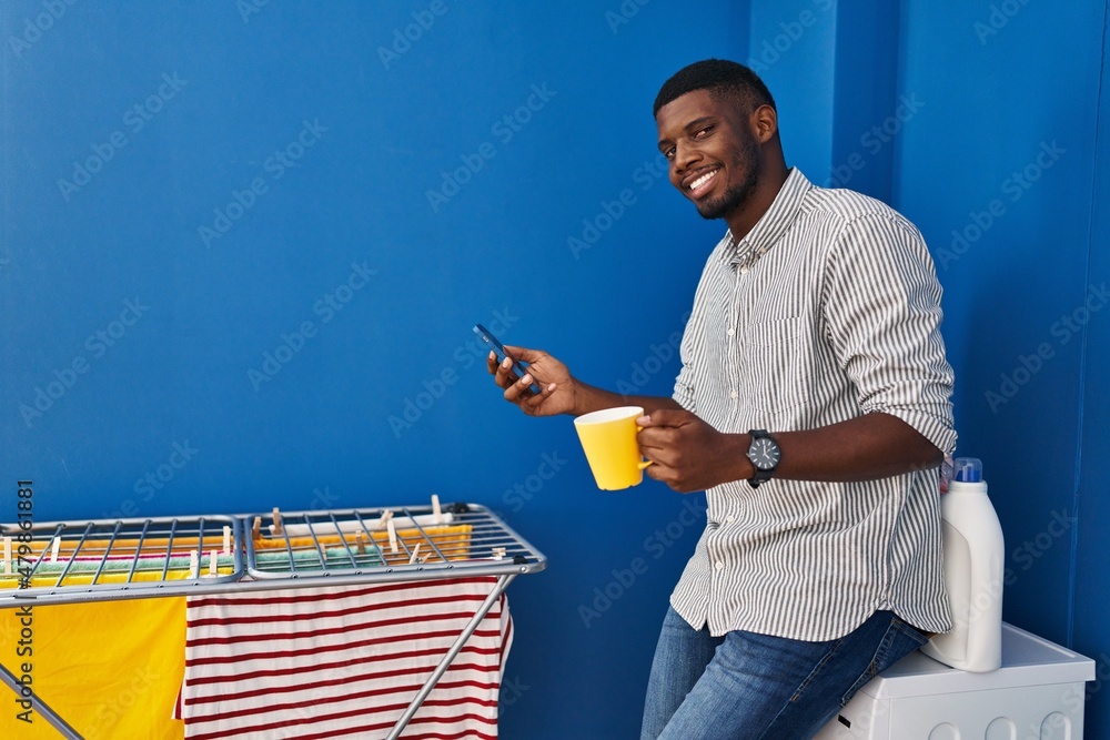 Young african american man using smartphone and drinking coffee waiting for washing machine at laundry room Young african american man using smartphone and drinking coffee waiting for washing machine at laundry room