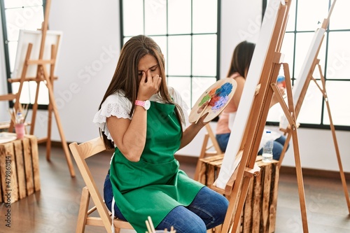 Young hispanic artist women painting on canvas at art studio tired rubbing nose and eyes feeling fatigue and headache. stress and frustration concept.