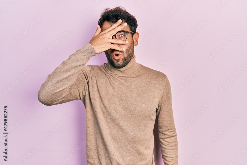 Handsome man with beard wearing turtleneck sweater and glasses peeking in shock covering face and eyes with hand, looking through fingers with embarrassed expression. Handsome man with beard wearing turtleneck sweater and glasses peeking in shock covering face and eyes with hand, looking through fingers with embarrassed expression.