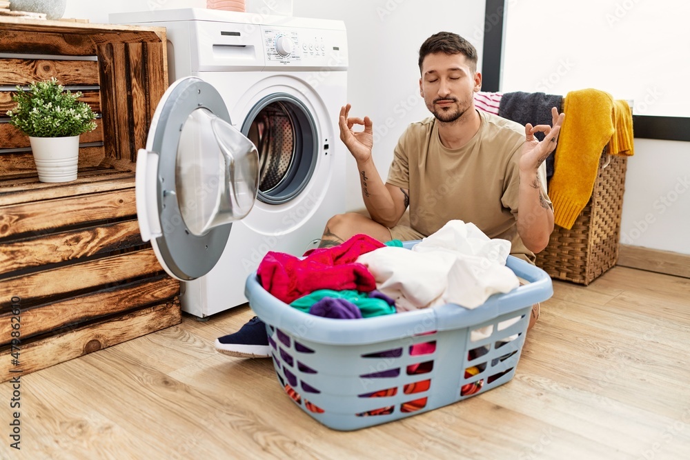 Young handsome man putting dirty laundry into washing machine relax and smiling with eyes closed doing meditation gesture with fingers. yoga concept. Young handsome man putting dirty laundry into washing machine relax and smiling with eyes closed doing meditation gesture with fingers. yoga concept.