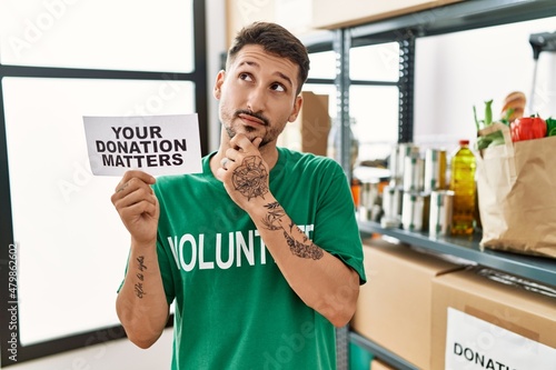 Young volunteer man holding your donation matters banner serious face thinking about question with hand on chin  thoughtful about confusing idea