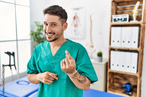 Young physiotherapist man working at pain recovery clinic beckoning come here gesture with hand inviting welcoming happy and smiling