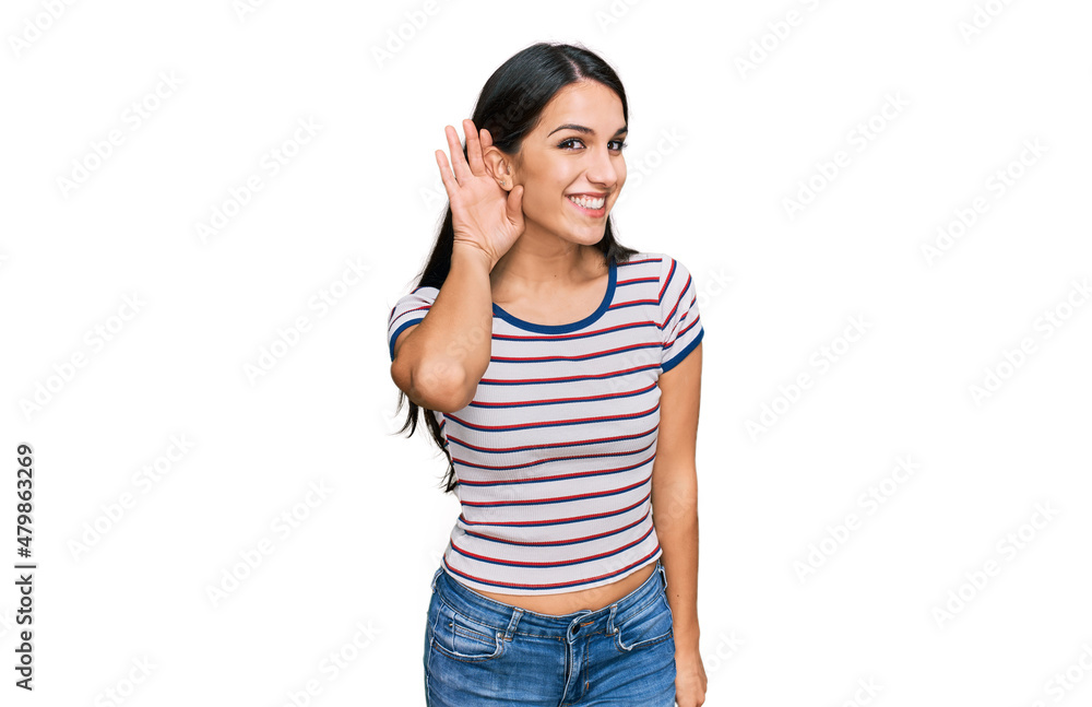Young hispanic girl wearing casual striped t shirt smiling with hand over ear listening an hearing to rumor or gossip. deafness concept. Young hispanic girl wearing casual striped t shirt smiling with hand over ear listening an hearing to rumor or gossip. deafness concept.