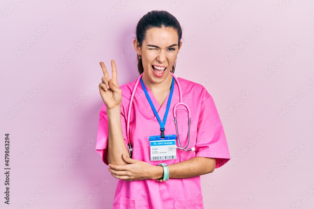 Young brunette woman wearing doctor uniform and stethoscope smiling with happy face winking at the camera doing victory sign with fingers. number two. Young brunette woman wearing doctor uniform and stethoscope smiling with happy face winking at the camera doing victory sign with fingers. number two.