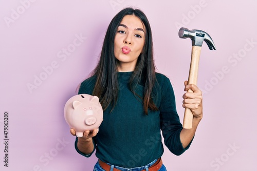 Young brunette woman holding piggy bank and hammer looking at the camera blowing a kiss being lovely and sexy. love expression.