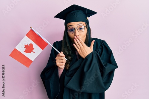 Young hispanic woman wearing graduation uniform holding canada flag covering mouth with hand, shocked and afraid for mistake. surprised expression