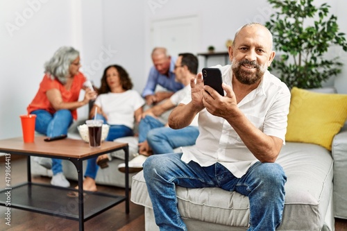 Group of middle age friends sitting on the sofa speaking. Man smiling happy using smartphone at home.