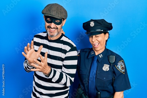 Middle age couple of hispanic woman and man wearing thief and police uniform winking looking at the camera with sexy expression, cheerful and happy face.