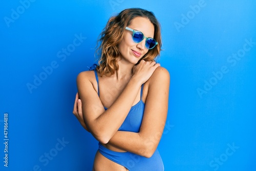 Young caucasian woman wearing swimsuit and sunglasses hugging oneself happy and positive, smiling confident. self love and self care