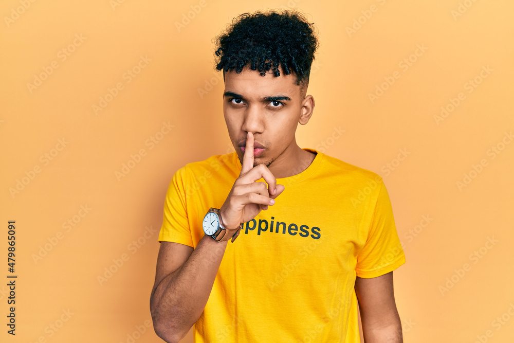 Young african american man wearing t shirt with happiness word message asking to be quiet with finger on lips. silence and secret concept. Young african american man wearing t shirt with happiness word message asking to be quiet with finger on lips. silence and secret concept.