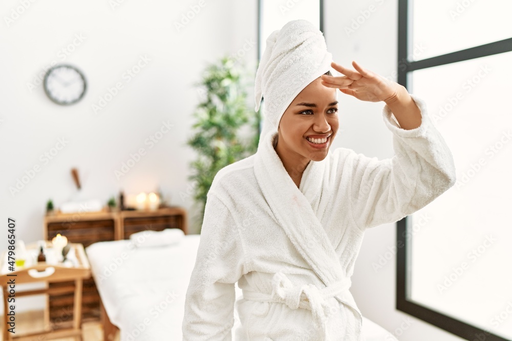 Young brunette woman wearing towel and bathrobe standing at beauty center very happy and smiling looking far away with hand over head. searching concept. Young brunette woman wearing towel and bathrobe standing at beauty center very happy and smiling looking far away with hand over head. searching concept.