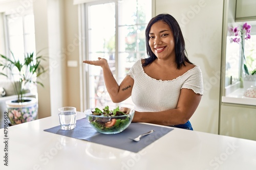 Young hispanic woman eating healthy salad at home smiling cheerful presenting and pointing with palm of hand looking at the camera.