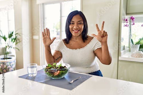 Young hispanic woman eating healthy salad at home showing and pointing up with fingers number eight while smiling confident and happy.