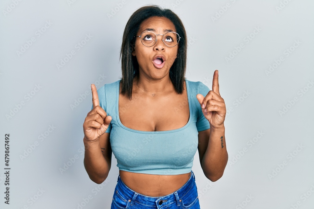 Young african american girl wearing casual clothes amazed and surprised looking up and pointing with fingers and raised arms. Young african american girl wearing casual clothes amazed and surprised looking up and pointing with fingers and raised arms.