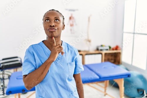 Black woman with braids working at pain recovery clinic thinking concentrated about doubt with finger on chin and looking up wondering