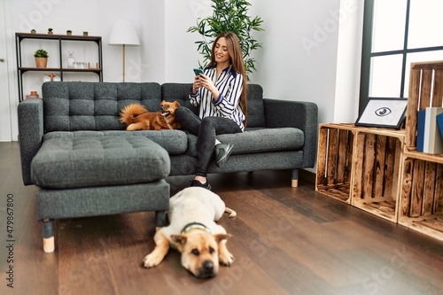 Young hispanic woman using smartphone sitting on sofa with dogs at home