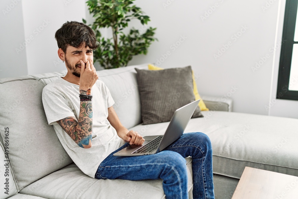 Hispanic man with beard sitting on the sofa smelling something stinky and disgusting, intolerable smell, holding breath with fingers on nose. bad smell Hispanic man with beard sitting on the sofa smelling something stinky and disgusting, intolerable smell, holding breath with fingers on nose. bad smell