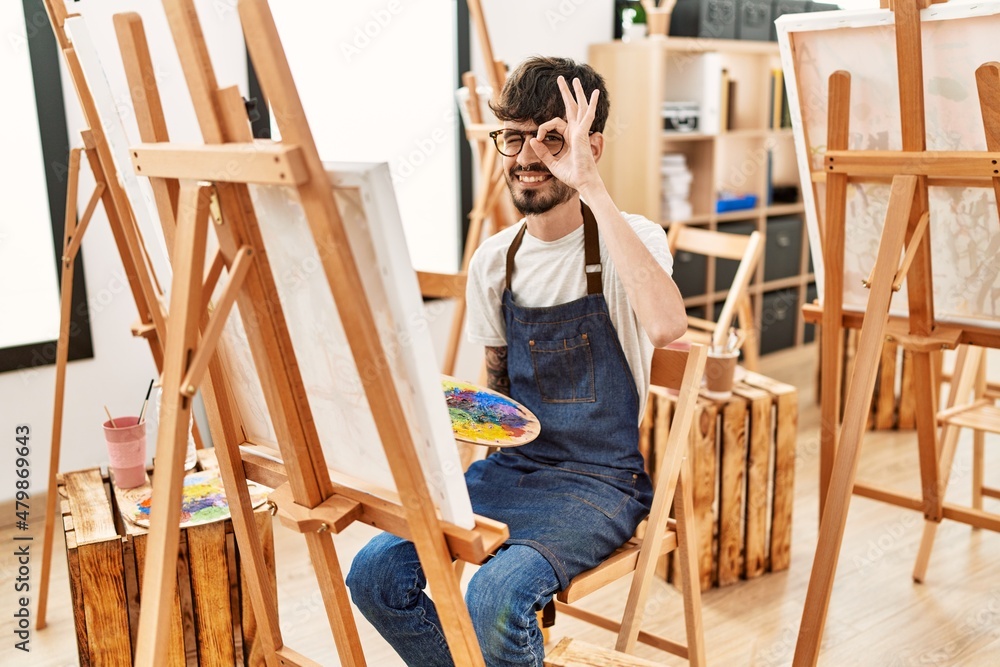 Hispanic man with beard at art studio smiling happy doing ok sign with hand on eye looking through fingers Hispanic man with beard at art studio smiling happy doing ok sign with hand on eye looking through fingers