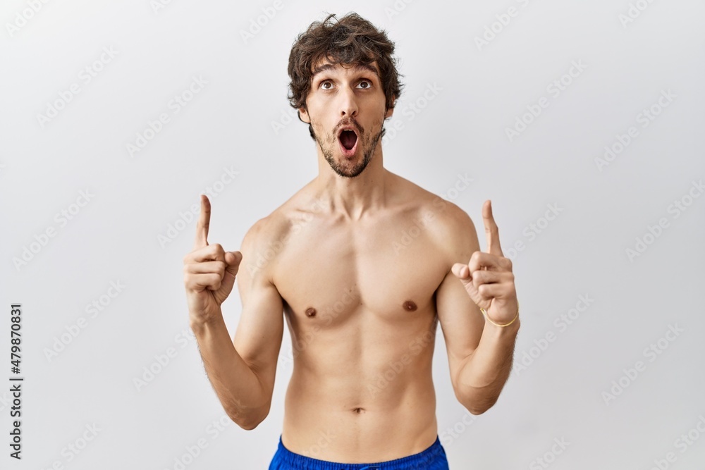 Young hispanic man standing shirtless over isolated, background amazed and surprised looking up and pointing with fingers and raised arms. Young hispanic man standing shirtless over isolated, background amazed and surprised looking up and pointing with fingers and raised arms.