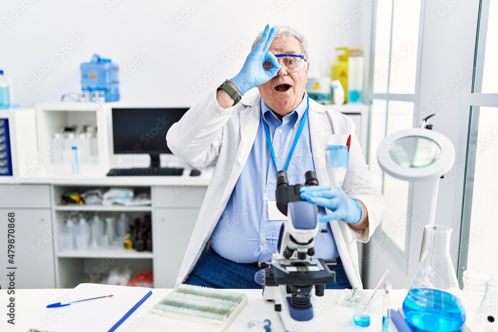 Senior caucasian man working at scientist laboratory doing ok gesture shocked with surprised face, eye looking through fingers. unbelieving expression. Senior caucasian man working at scientist laboratory doing ok gesture shocked with surprised face, eye looking through fingers. unbelieving expression.