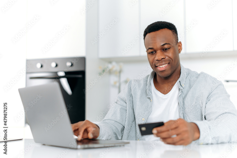 Joyful black man sits on kitchen at home, dressed in casual wear, using laptop and credit card for online shopping, is entering data for payment, online transaction, order home delivery, smiles Joyful black man sits on kitchen at home, dressed in casual wear, using laptop and credit card for online shopping, is entering data for payment, online transaction, order home delivery, smiles