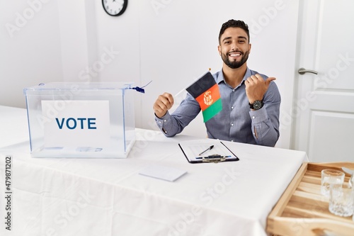 Young handsome man with beard at political campaign election holding afghanistan flag pointing thumb up to the side smiling happy with open mouth