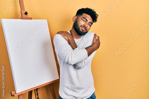 Arab man with beard standing by painter easel stand hugging oneself happy and positive, smiling confident Fototapet