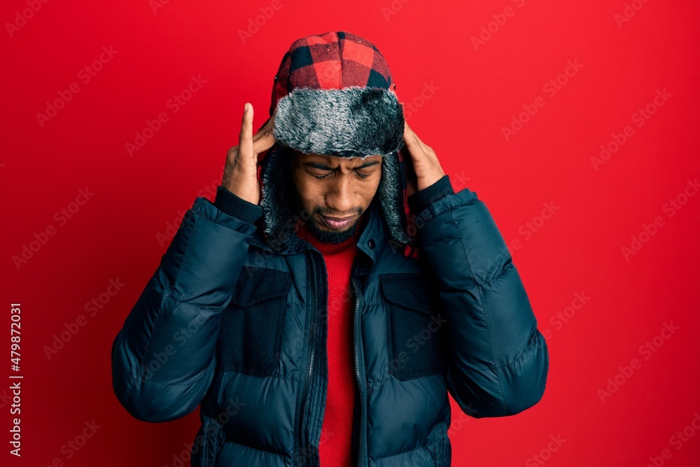 Young african american man with beard wearing winter hat and coat with hand on head for pain in head because stress. suffering migraine.
