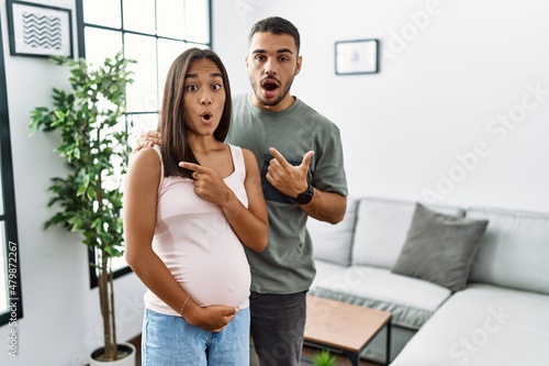 Young interracial couple expecting a baby, touching pregnant belly surprised pointing with finger to the side, open mouth amazed expression.
