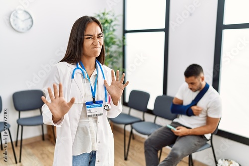 Young asian doctor woman at waiting room with a man with a broken arm disgusted expression  displeased and fearful doing disgust face because aversion reaction. with hands raised