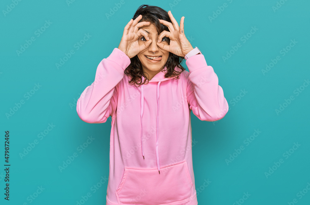 Young hispanic woman wearing casual sweatshirt doing ok gesture like binoculars sticking tongue out, eyes looking through fingers. crazy expression. Young hispanic woman wearing casual sweatshirt doing ok gesture like binoculars sticking tongue out, eyes looking through fingers. crazy expression.