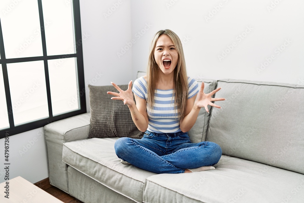 Asian young woman sitting on the sofa at home crazy and mad shouting and yelling with aggressive expression and arms raised. frustration concept. Asian young woman sitting on the sofa at home crazy and mad shouting and yelling with aggressive expression and arms raised. frustration concept.