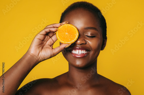 Portrait of an african girl covering eye with orange slice isolated over orange color background. Spa body care. Beauty fashion model. Beauty face. Beauty