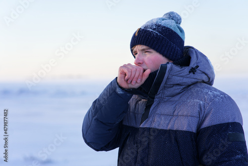 Fotografie Sick ill guy, young man in hat coughing in his fist, hand, suffering from cough, bronchitis at cold snowy frosty winter day outdoors