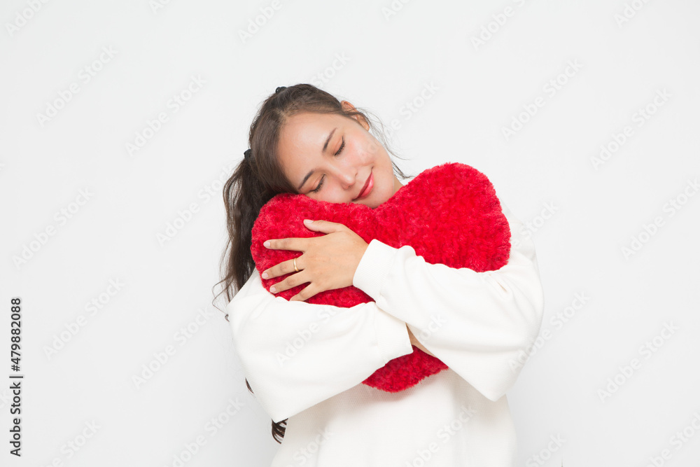 Beautiful Asian woman wearing white sweater closing her eyes and hugging red heart shape pillow on white background and copy space for Valentine Day concept.
