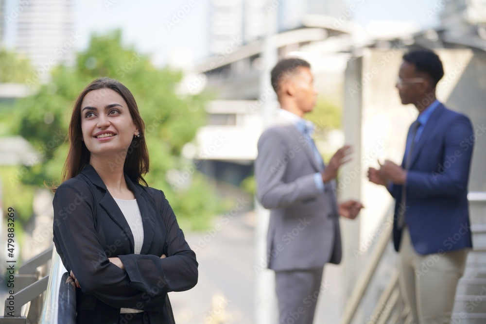 A confident businesswoman smiles while standing with her arms crossed, with two colleagues conversing in the background, showcasing professionalism and a collaborative work environment. A confident businesswoman smiles while standing with her arms crossed, with two colleagues conversing in the background, showcasing professionalism and a collaborative work environment.