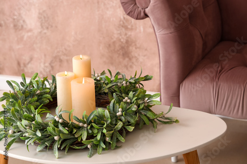 Burning candles with wreath on table near pink armchair, closeup