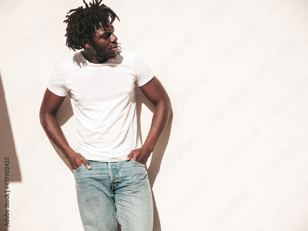 Portrait of handsome hipster model.Unshaven African man dressed in white summer t-shirt and jeans. Fashion male with dreadlocks hairstyle posing near white wall in the street Portrait of handsome hipster model.Unshaven African man dressed in white summer t-shirt and jeans. Fashion male with dreadlocks hairstyle posing near white wall in the street