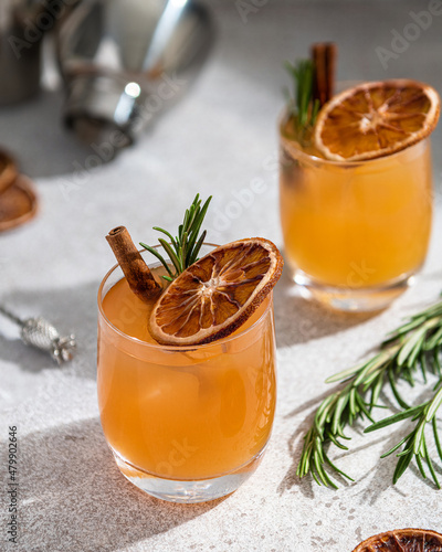 Alcoholic cocktail decorated with rosemary sprig, cinnamon stick and dried orange slice on light background