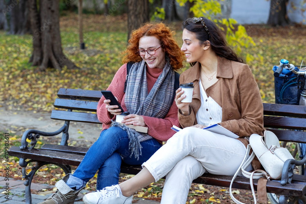 Two woman sitting at the bench in public park with coffee to go on hands and talk Two woman sitting at the bench in public park with coffee to go on hands and talk