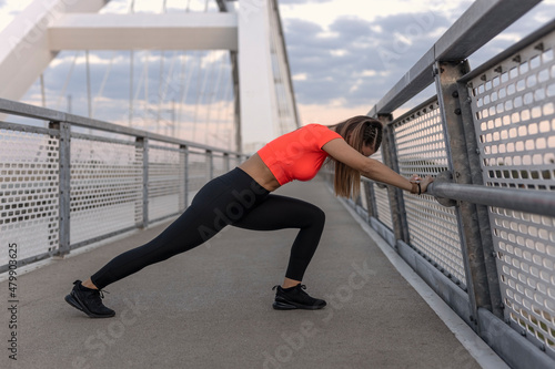 Young happy focused fitness girl in black yoga pants and orange short shirt work out and stretch her body on the bridge footpath during the day. Front view.