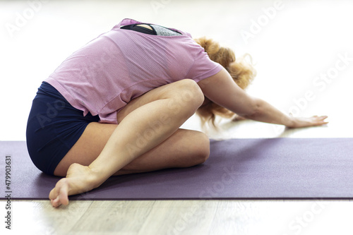 Middle aged slim blond woman in purple sportswear stretching lower back on floor mat in yoga studio. Side view of back flexibility pose.