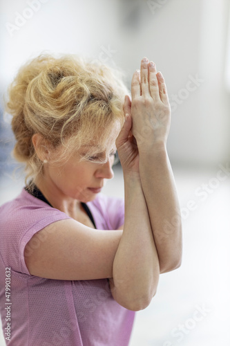 Middle aged slim blond woman in purple sportswear makes garudasana yoga pose with hands in yoga studio. Front view. photo