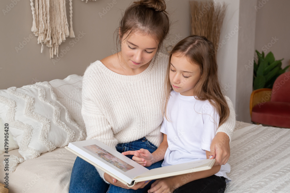 mother and daughter are sitting at home on the bed flipping through and looking at a book with photos. important moments of life in a photo album. Cute sisters read an interesting fairy tale together mother and daughter are sitting at home on the bed flipping through and looking at a book with photos. important moments of life in a photo album. Cute sisters read an interesting fairy tale together