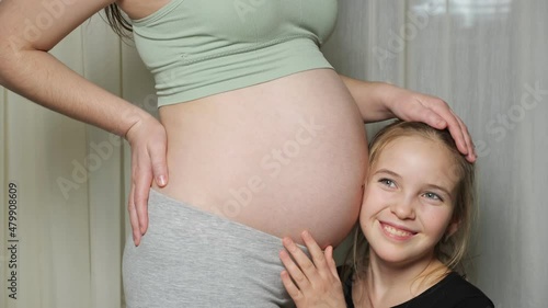 Elder blonde-haired teen daughter listens to baby moves in mother pregnant bare belly and hugs woman smiling with loving atmosphere closeup. photo