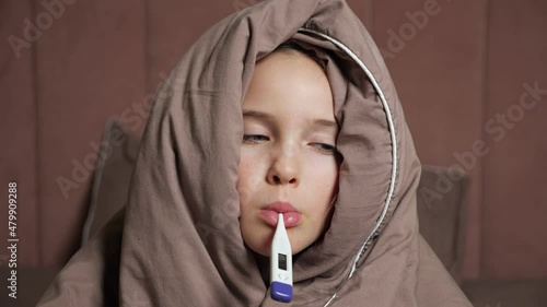 Sick teen girl holds electronic thermometer in mouth measuring temperature covering head with warm brown blanket in bedroom extreme closeup. photo