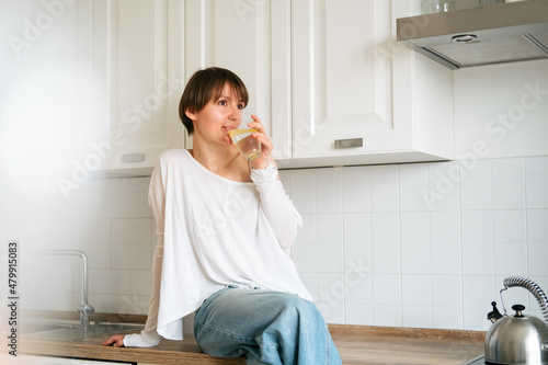 Young beautiful woman in casual clothes is sitting on countertop in kitchen and enjoying new day. Wellbeing, female health and good morning concept.